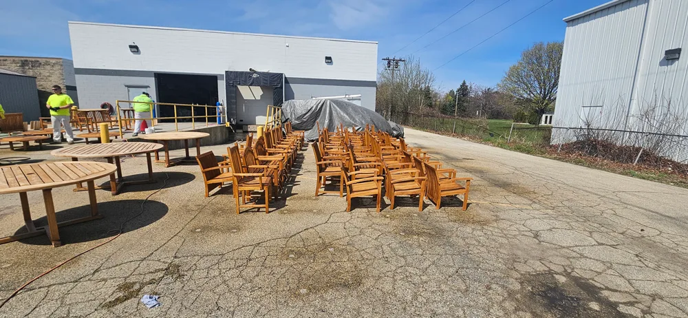 Outdoor scene behind an industrial building showing rows of wooden patio chairs and round tables arranged on a cracked asphalt surface, with workers in high-visibility clothing nearby and a tarp-covered object in the background.
