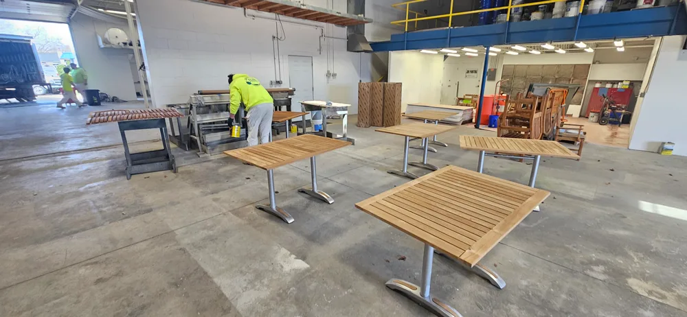 Outdoor industrial area with rows of wooden patio chairs neatly lined up on cracked pavement, several round wooden tables to the left, and workers in high-visibility clothing near a building with open garage doors and a tarp-covered object.