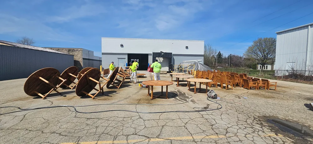 Outdoor industrial area with rows of wooden patio chairs neatly lined up on cracked pavement, several round wooden tables to the left, and workers in high-visibility clothing near a building with open garage doors and a tarp-covered object.
